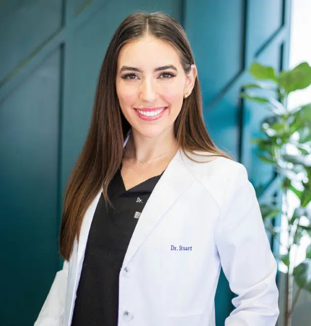 Female dentist in white coat smiling at camera in modern dental office setting.